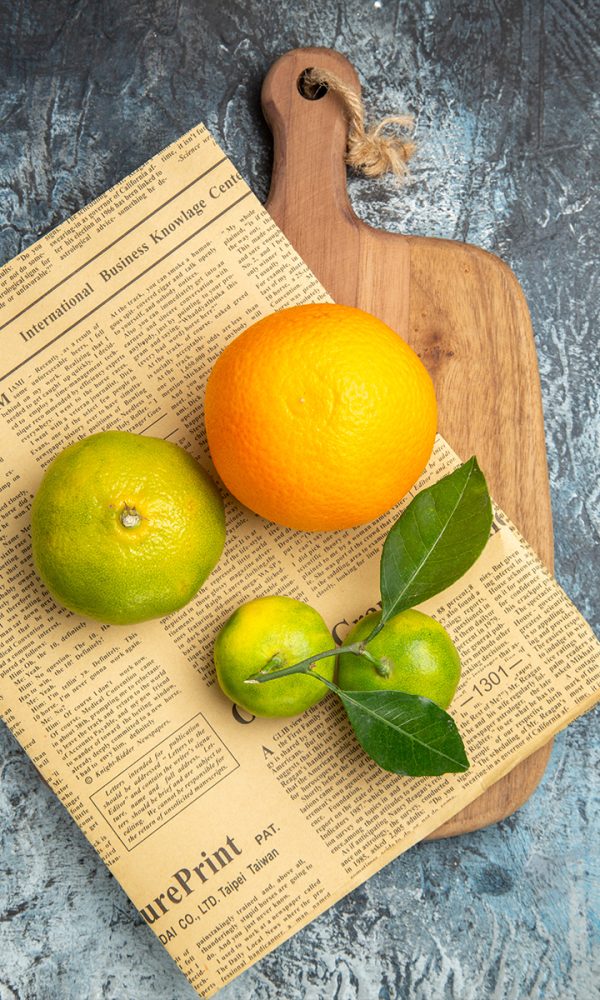 Overhead view fresh citrus fruits with leaves newpaper wooden cutting board cut half forms gray table 600x1000
