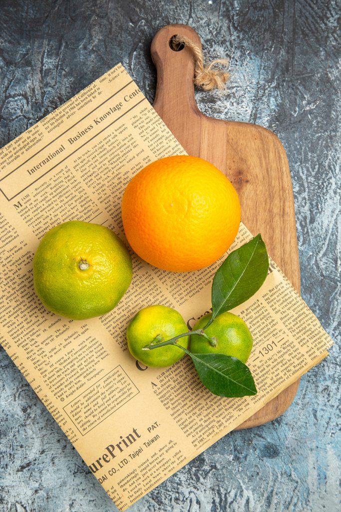 Overhead view fresh citrus fruits with leaves newpaper wooden cutting board cut half forms gray table 683x1024